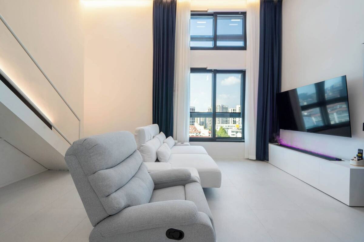 A modern living room with a grey sectional sofa, a flat-screen TV mounted on the wall, and a large window with a city view.