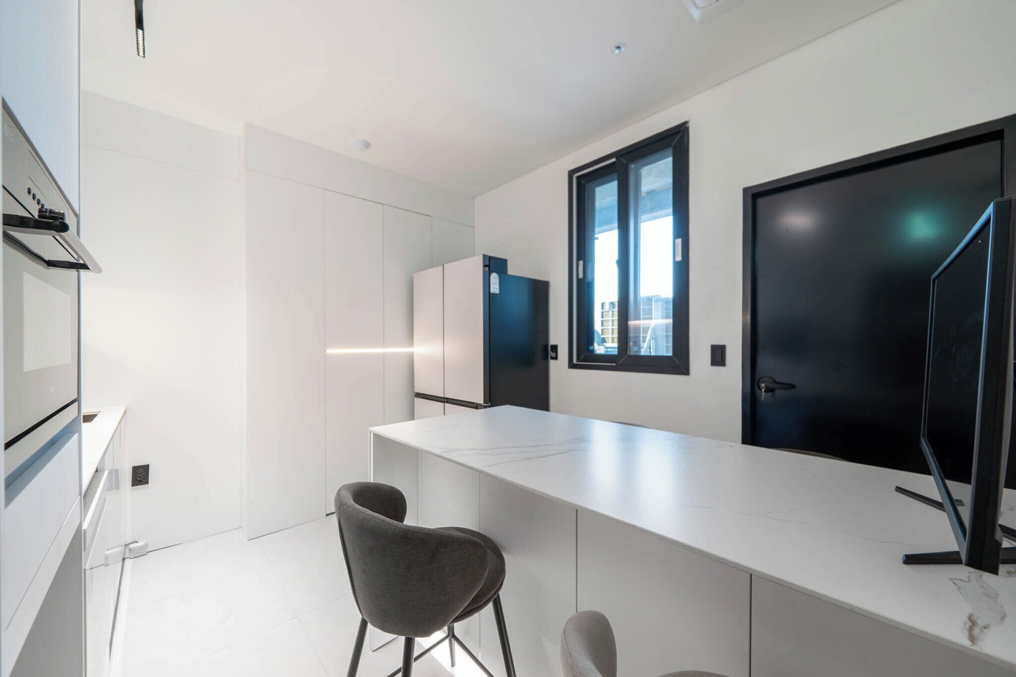A modern kitchen with white cabinetry, a marble countertop, two grey chairs, and a black-framed TV.