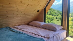 A bedroom with a sloped ceiling, wooden walls, a bed with pink bedding, and a view of a green landscape through a large window. - Svanieri cozy cottage in the mountains (Mestia Municipality)