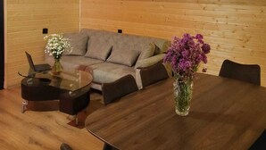 A modern living room with a wooden ceiling, a grey sofa, a glass coffee table, a wooden dining table, and two vases with flowers. - Svanieri cozy cottage in the mountains (Mestia Municipality)