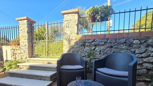 Un área de estar al aire libre con dos sillas negras y una mesa redonda, un muro de piedra y una reja metálica al fondo.