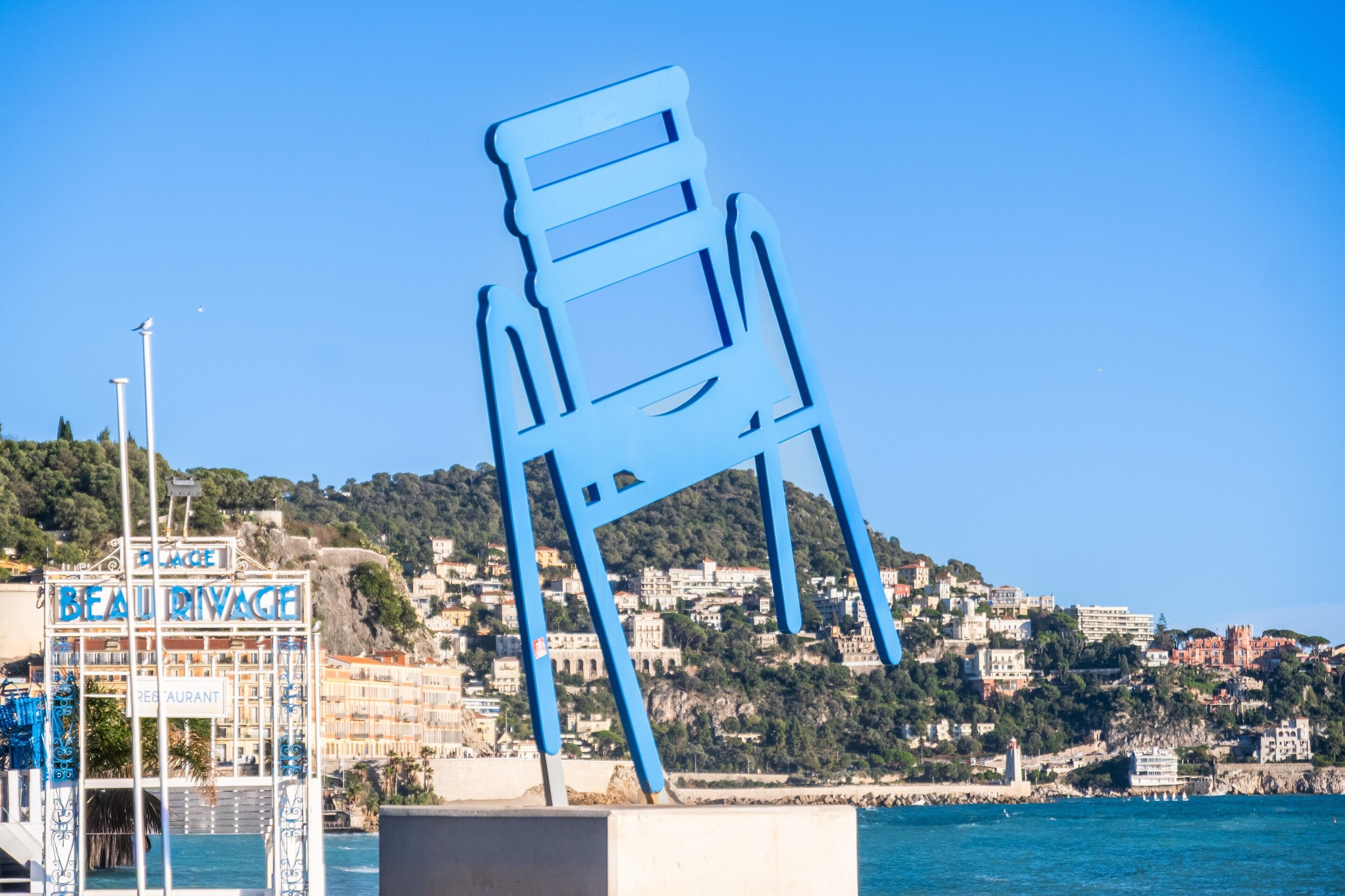 A large blue chair sculpture in front of a coastal town with buildings and a sign that reads 'BEAU RIVAGE'.