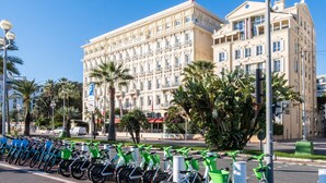 Een groot hotel in klassieke stijl, met een rij fietsen en palmbomen op de voorgrond.