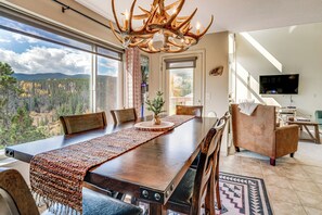 Un comedor con una gran mesa de madera, sillas y una araña. Se ve un bosque a través de amplias ventanas.