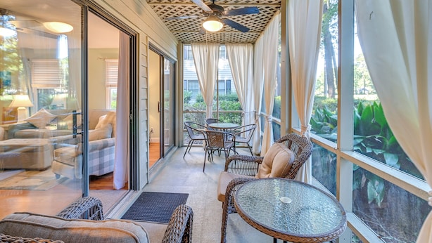 Une terrasse ensoleillée, meublée de mobilier en osier, dotée d’un ventilateur de plafond et d’une porte vitrée donnant sur un espace de vie intérieur.