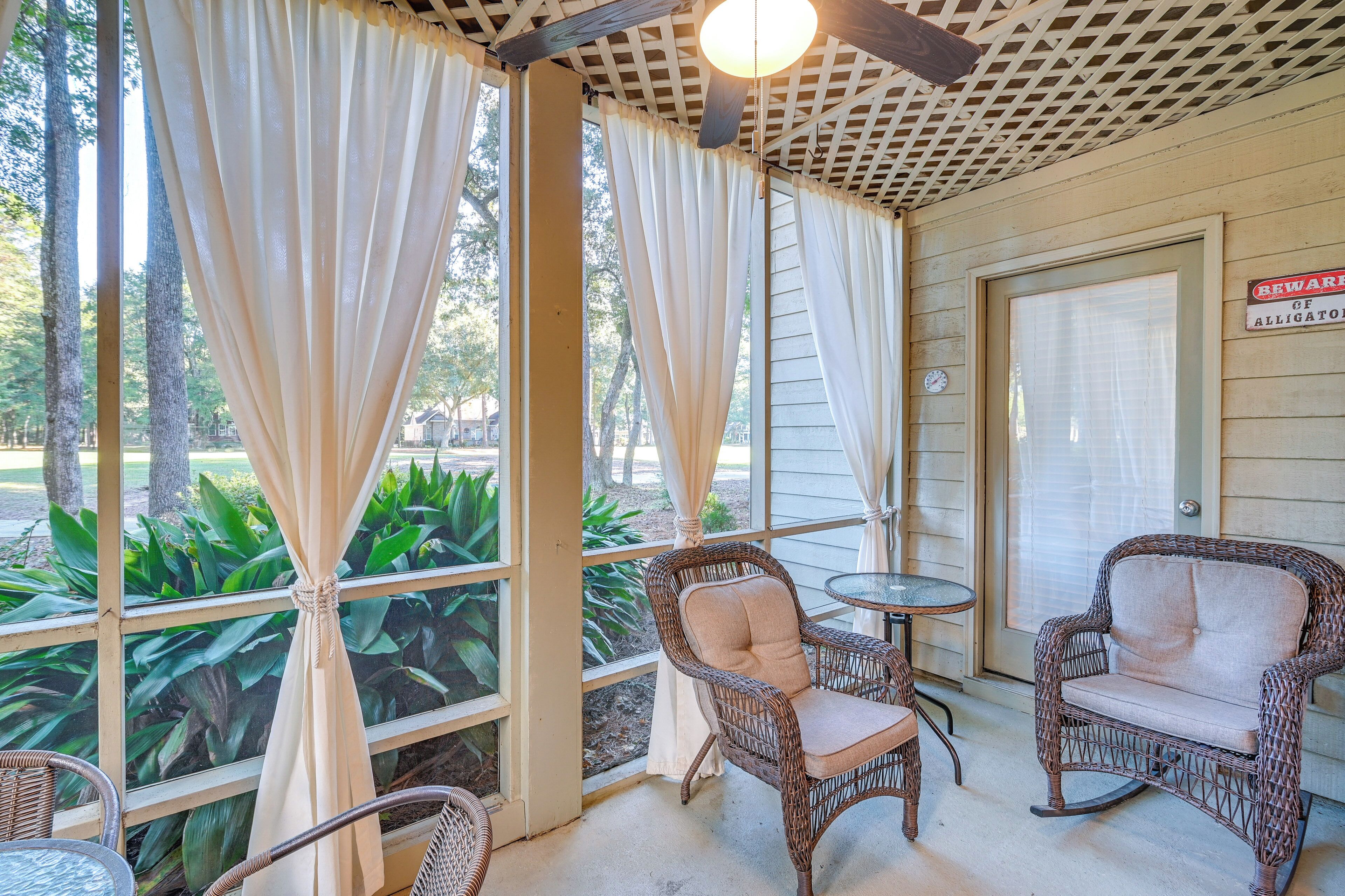 A covered porch with wicker chairs and a table, a sign warning of alligators, and a view of greenery outside.