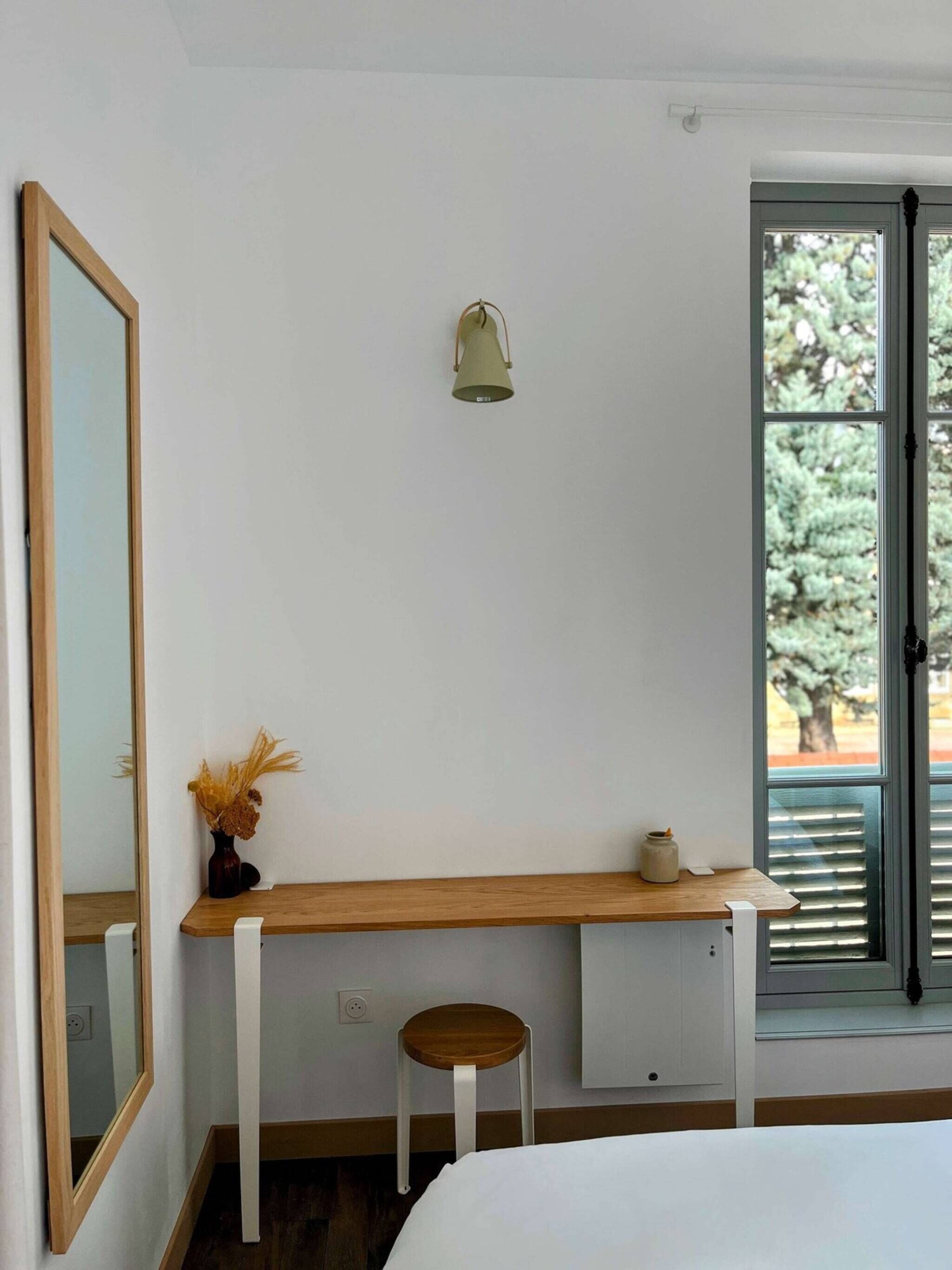 Un dormitorio con un escritorio de madera, un taburete pequeño, una mesita de noche con un florero, una lámpara de pared y una ventana con persianas.