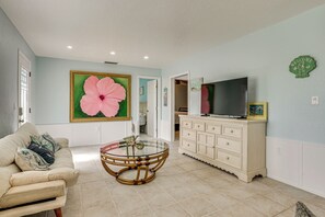 A living room with a round glass coffee table, a beige sofa, a large floral painting, a television on a cabinet, and a "WELCOME" sign on the wall. - Coastal Escape w/ Yard Near Anna Maria Island (Bradenton)