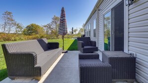 Eine überdachte Terrasse mit Korbmöbeln, einem gestreiften Sonnenschirm und Blick auf eine grüne Rasenfläche und Bäume.