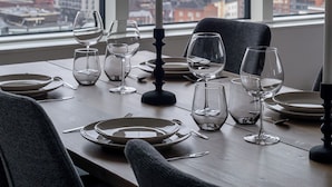 A dining table set with wine glasses and plates, two pendant lights, and a candle, with a cityscape view through large windows. - 12th Floor Sauna Retreat with Views (Tampere)