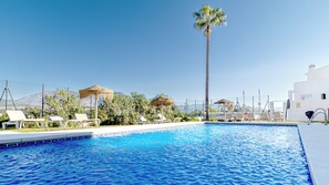 En swimmingpool med liggestole og palmer, omgivet af et hegn og hvide bygninger.