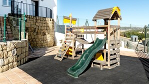 A playground with a slide and climbing structures, set against a white building with a stone wall and a metal fence. - Beautiful apartment, 10 minutes walk from the beach (Urbanización Estrella de la Bahía)
