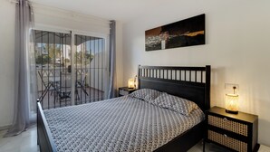 A bedroom with a bed, a nightstand, a lamp, a chair, and a view of the outside. - Beautiful apartment, 10 minutes walk from the beach (Urbanización Estrella de la Bahía)