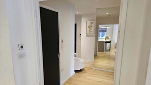 En hall med trägolv, vita väggar och en svart dörr. Längre bort står en vit stol, och en spegel reflekterar en del av rummet.