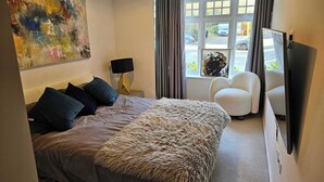 Ett modernt sovrum med en säng, en mjuk pläd, ett skrivbord med en lampa, en tv och ett fönster med gardiner.