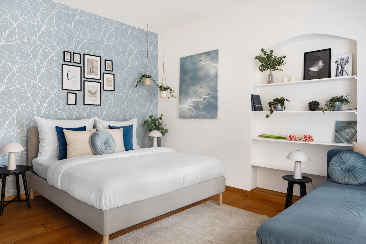 A modern bedroom with a bed, bedside tables, a wall with framed pictures, and shelves with plants and decorative items.
