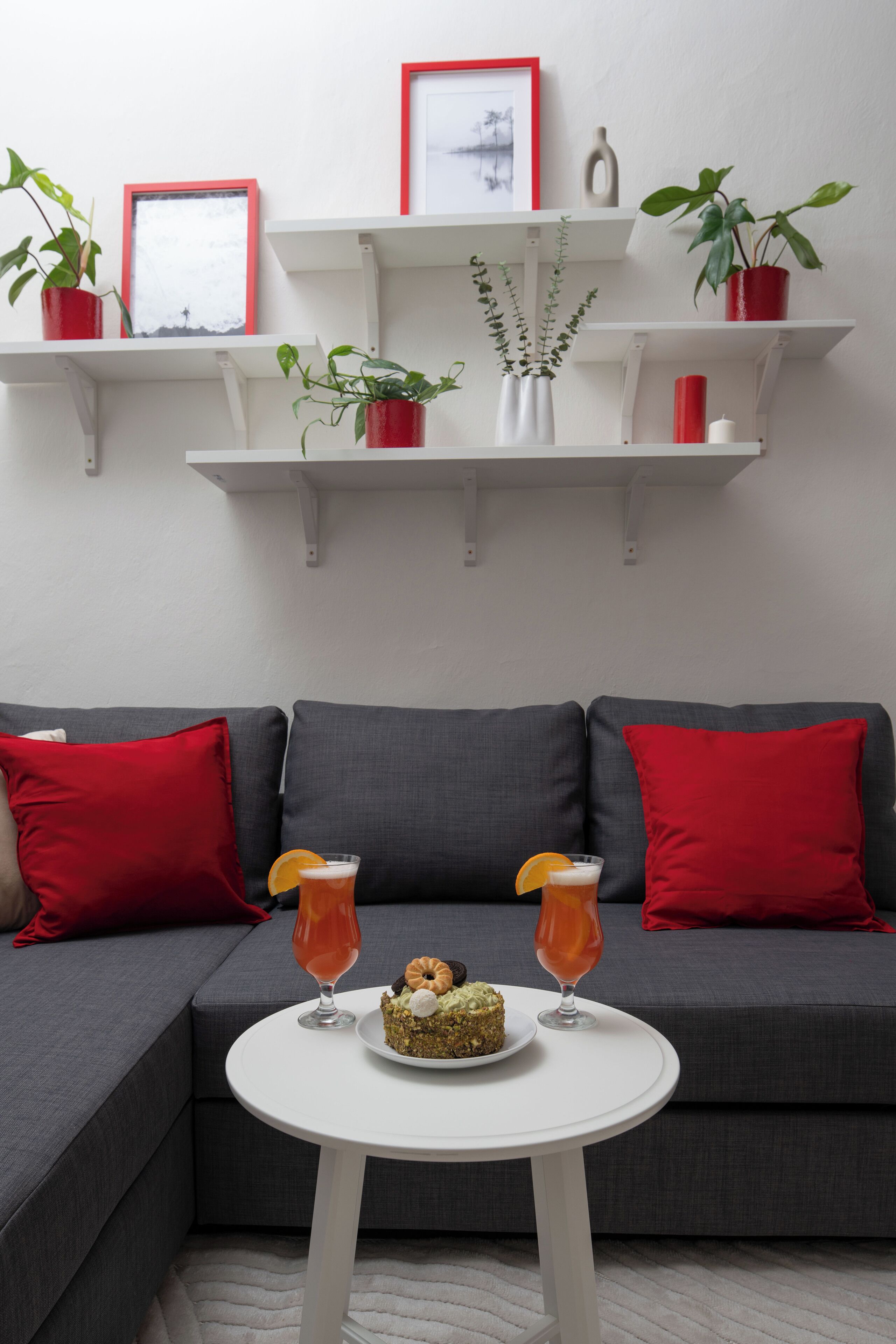 A modern living room with a grey sectional sofa, red cushions, a white round table with a dessert and drinks, and white shelves with plants and decorative items.