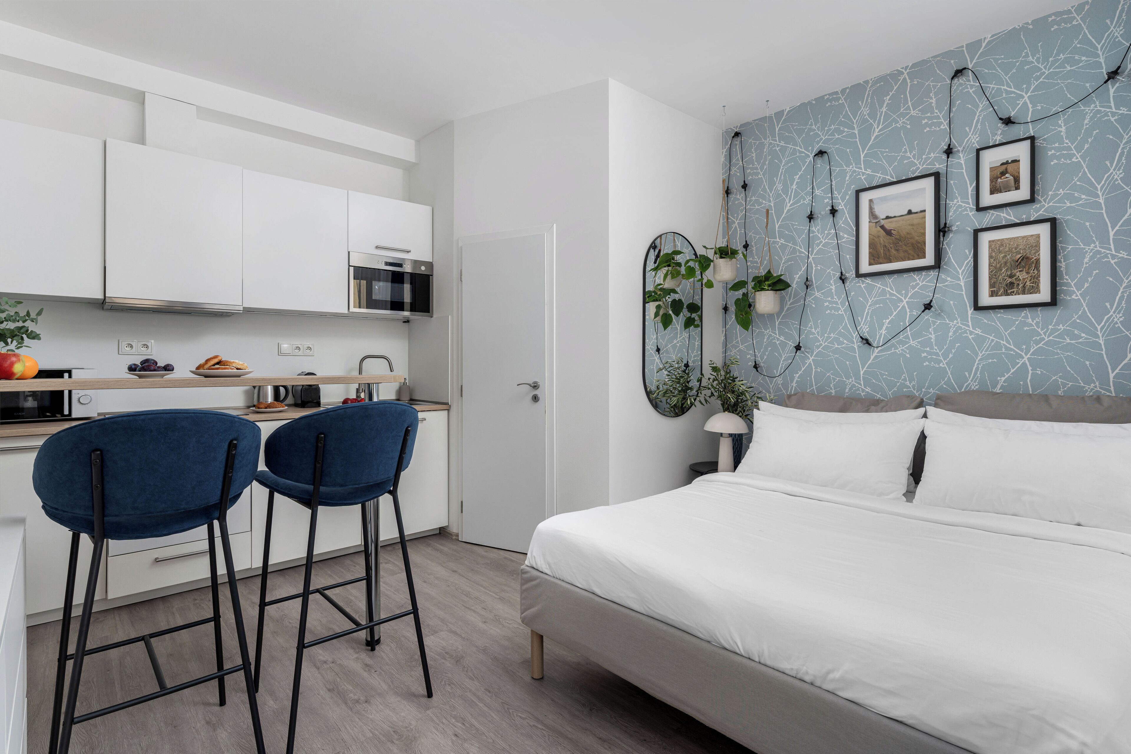 Ein modernes Schlafzimmer mit einem großen Bett, einer kleinen Küchenzeile mit weißen Schränken sowie einer blauen Wand mit gerahmten Bildern und Pflanzen.