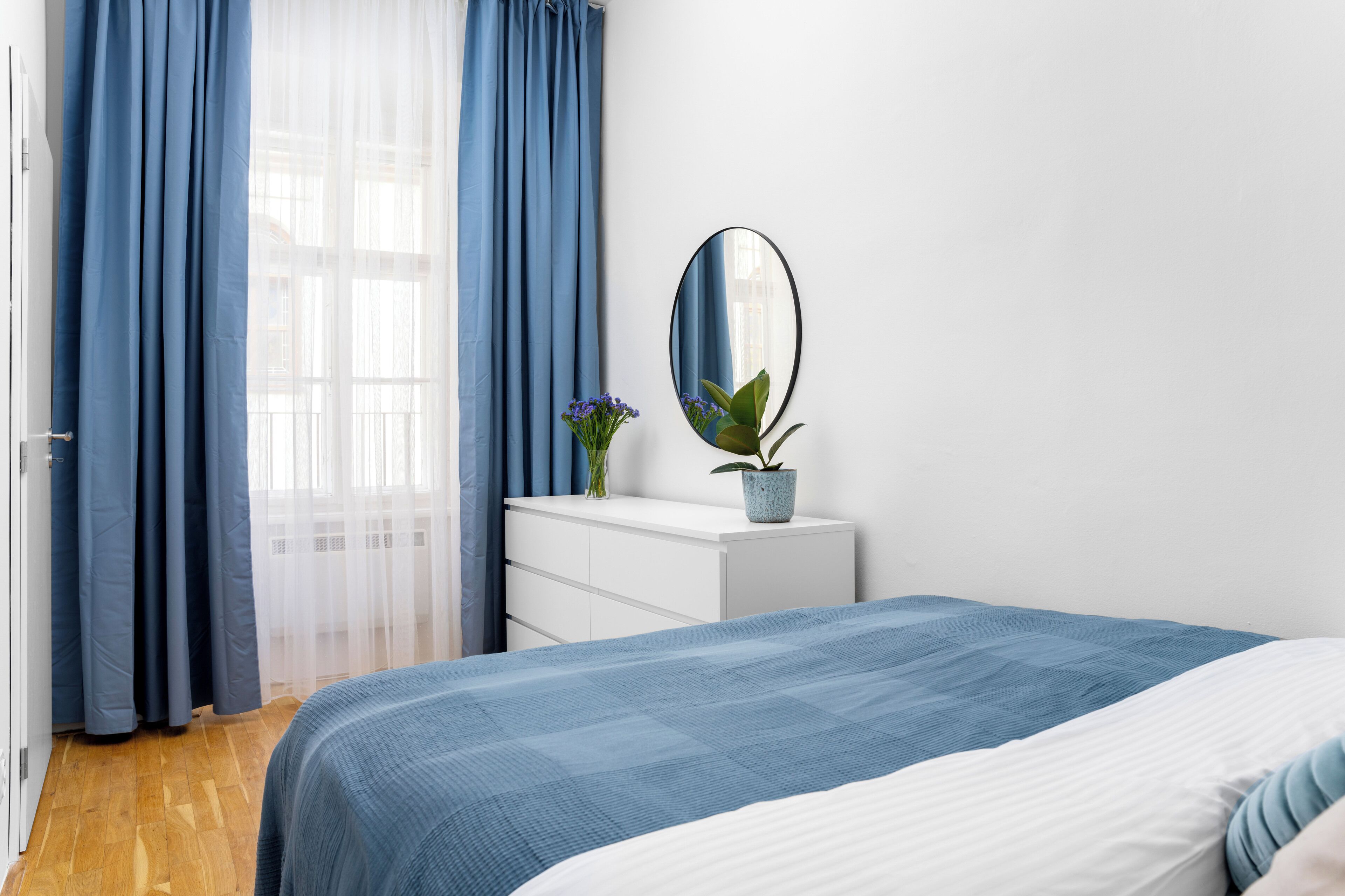 Une chambre à coucher avec un lit, une commode, un miroir et une fenêtre avec des rideaux bleus.