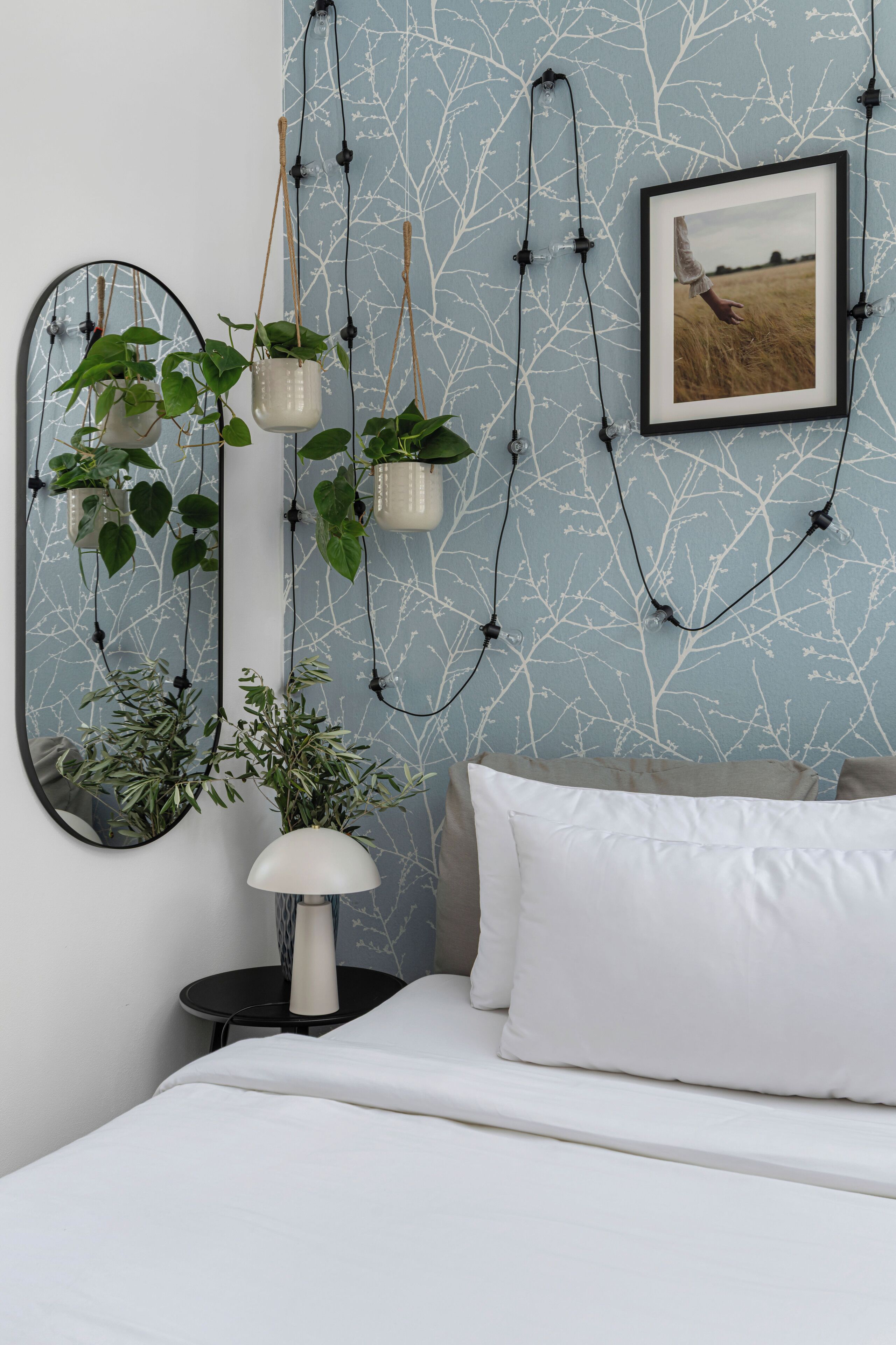 A bedroom with a white bed, a round mirror, and a wall-mounted picture. There are hanging plants and a small table with a lamp.