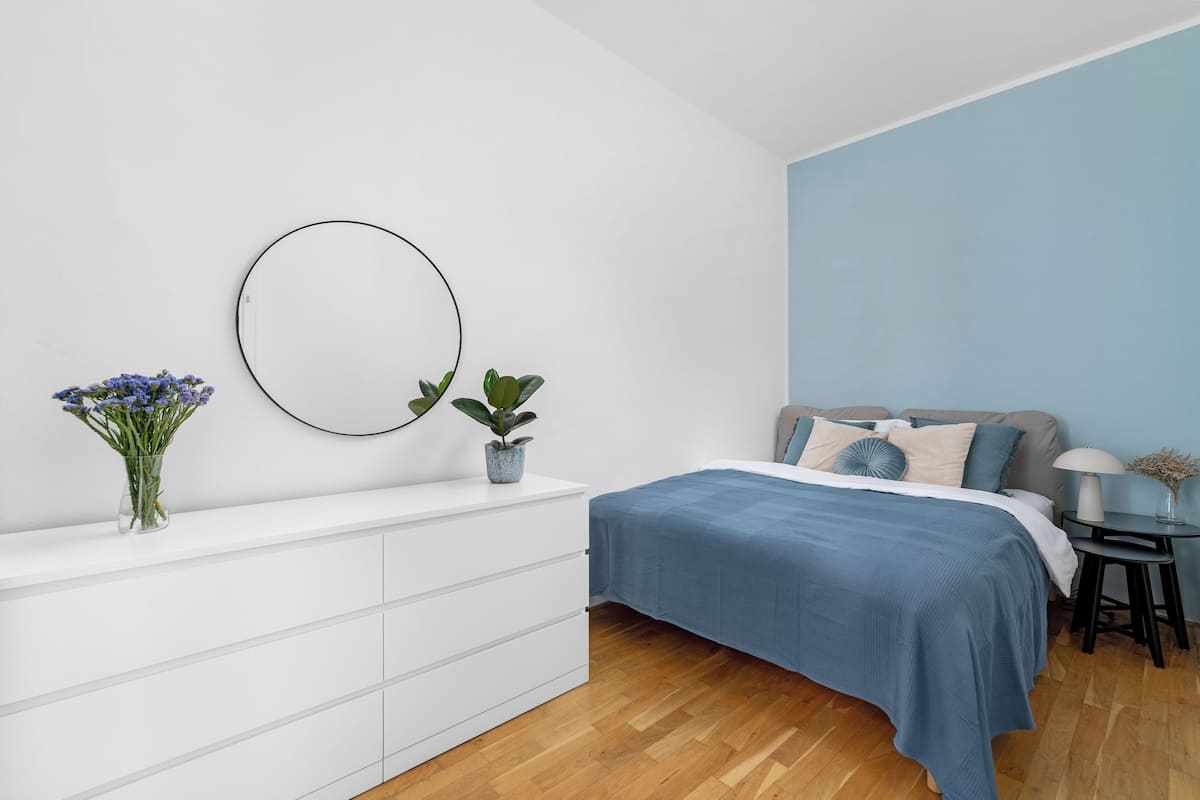 A bedroom with a bed, a dresser, a mirror, and a small table with a lamp.