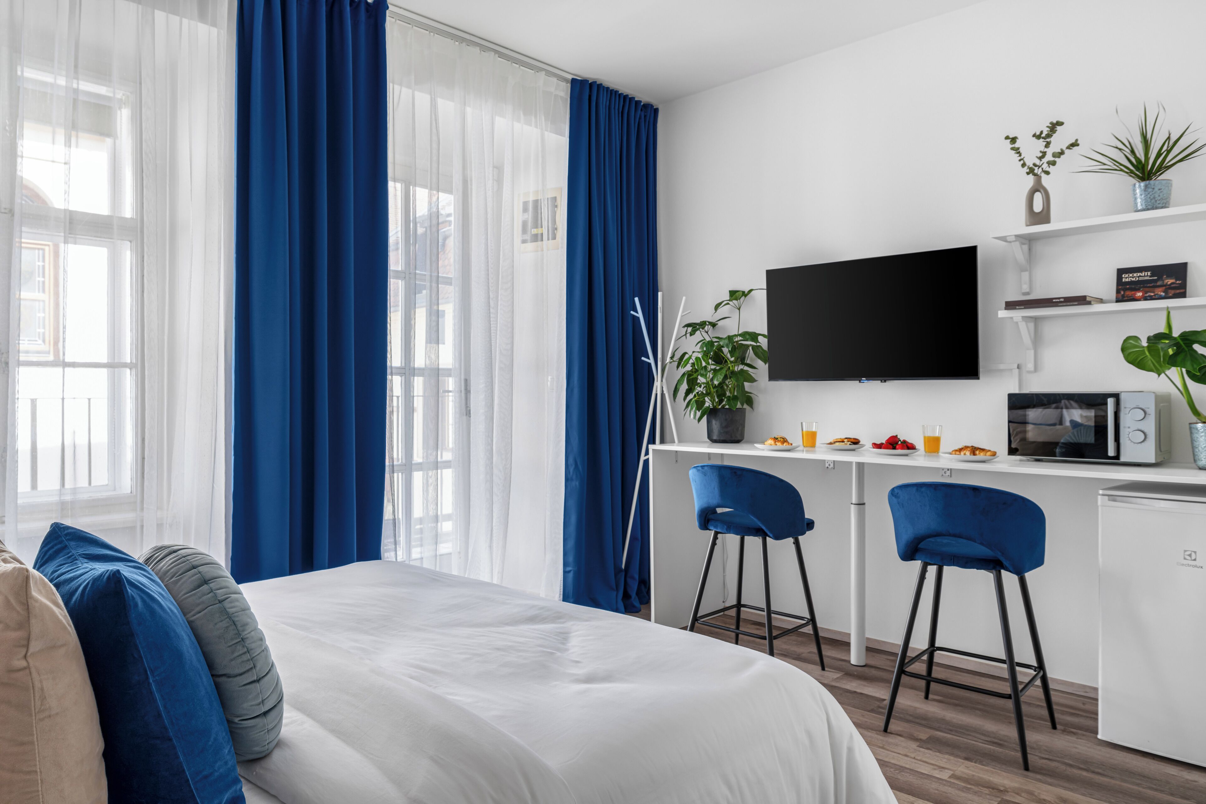 A modern hotel room with a bed, a desk with a computer, a TV, and a kitchenette.