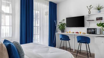 A modern hotel room with a bed, a desk with a computer, a TV, and a kitchenette.