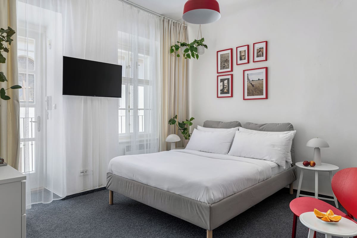 A hotel room with a bed, a TV mounted on the wall, a red chair, and a small table with fruit.
