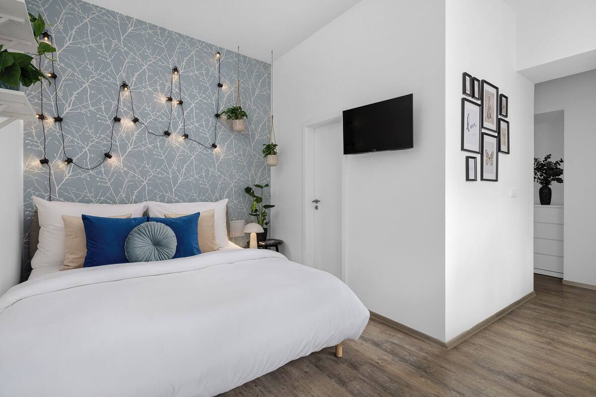 A modern bedroom with a white bed, blue and beige pillows, a marble-patterned wall, hanging plants, a TV, and framed pictures.