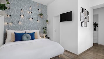 Une chambre moderne avec un lit blanc, des coussins bleus et beiges, un mur à motifs de marbre, des plantes suspendues, un téléviseur et des tableaux encadrés.