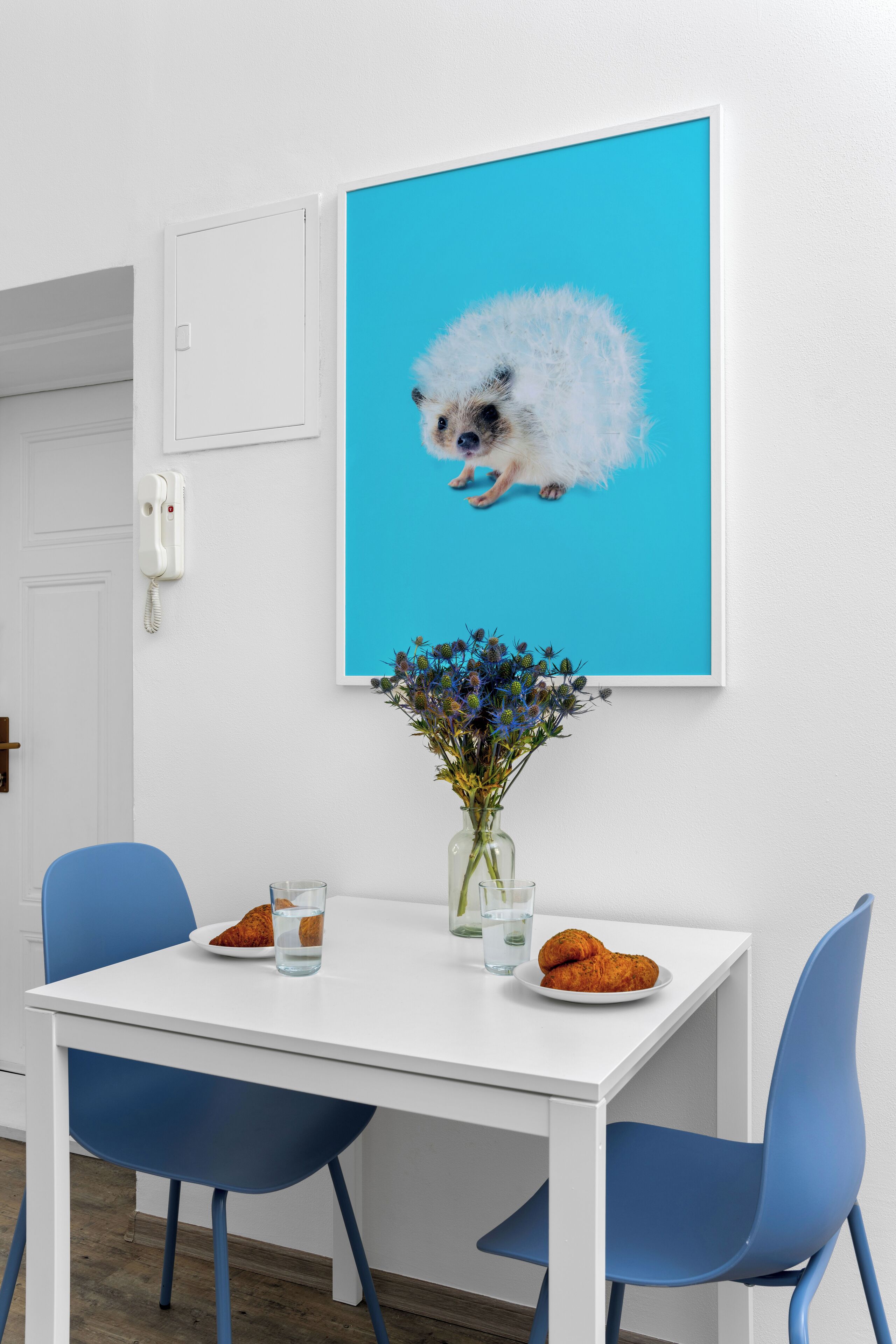 Un comedor con una mesa blanca, sillas azules, un cuadro con un erizo, un florero con flores y un plato de cruasanes.