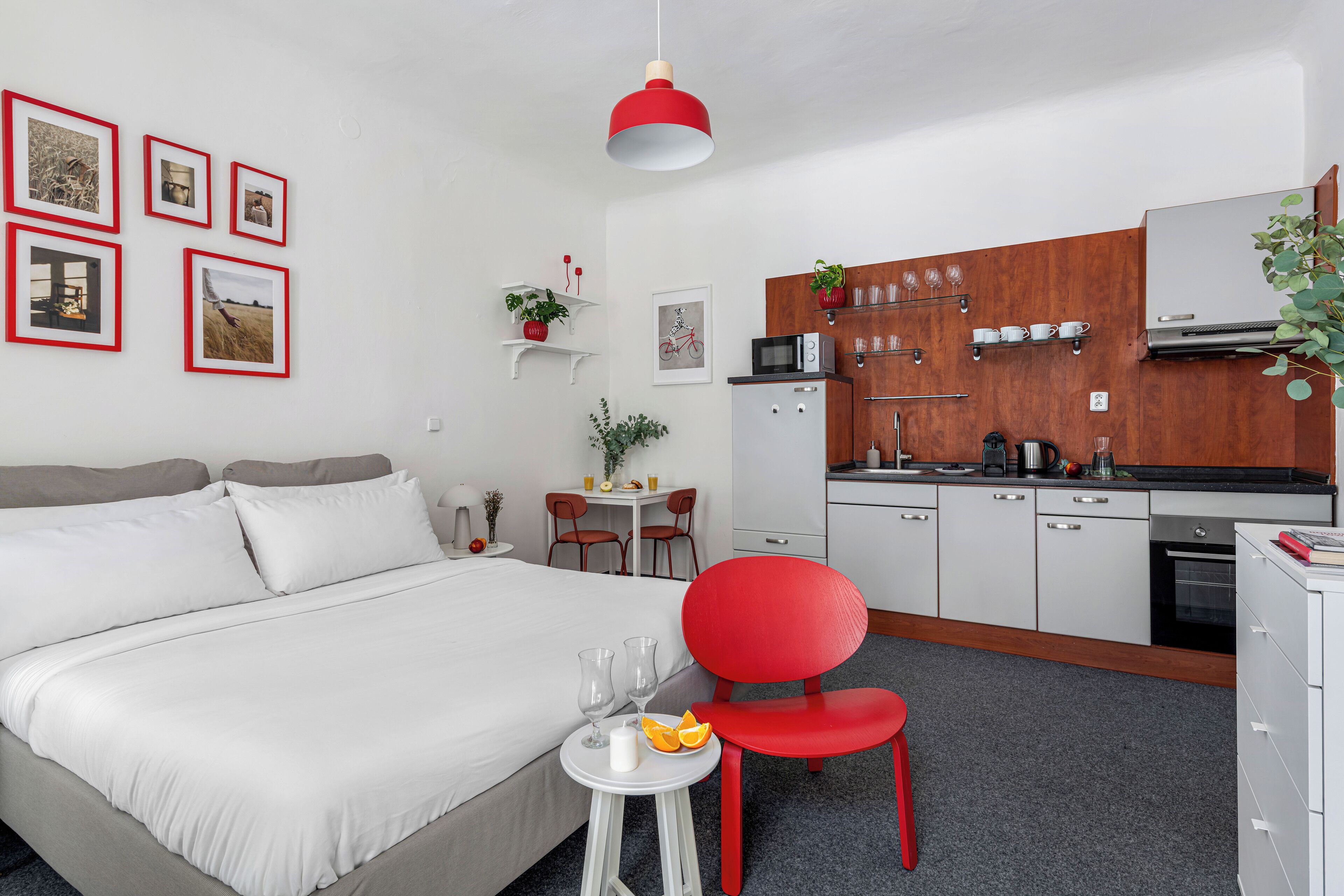 Ein modernes Hotelzimmer mit einem Bett, einem roten Sessel, einem kleinen Tisch mit Gläsern und Früchten sowie einer Küchenzeile.