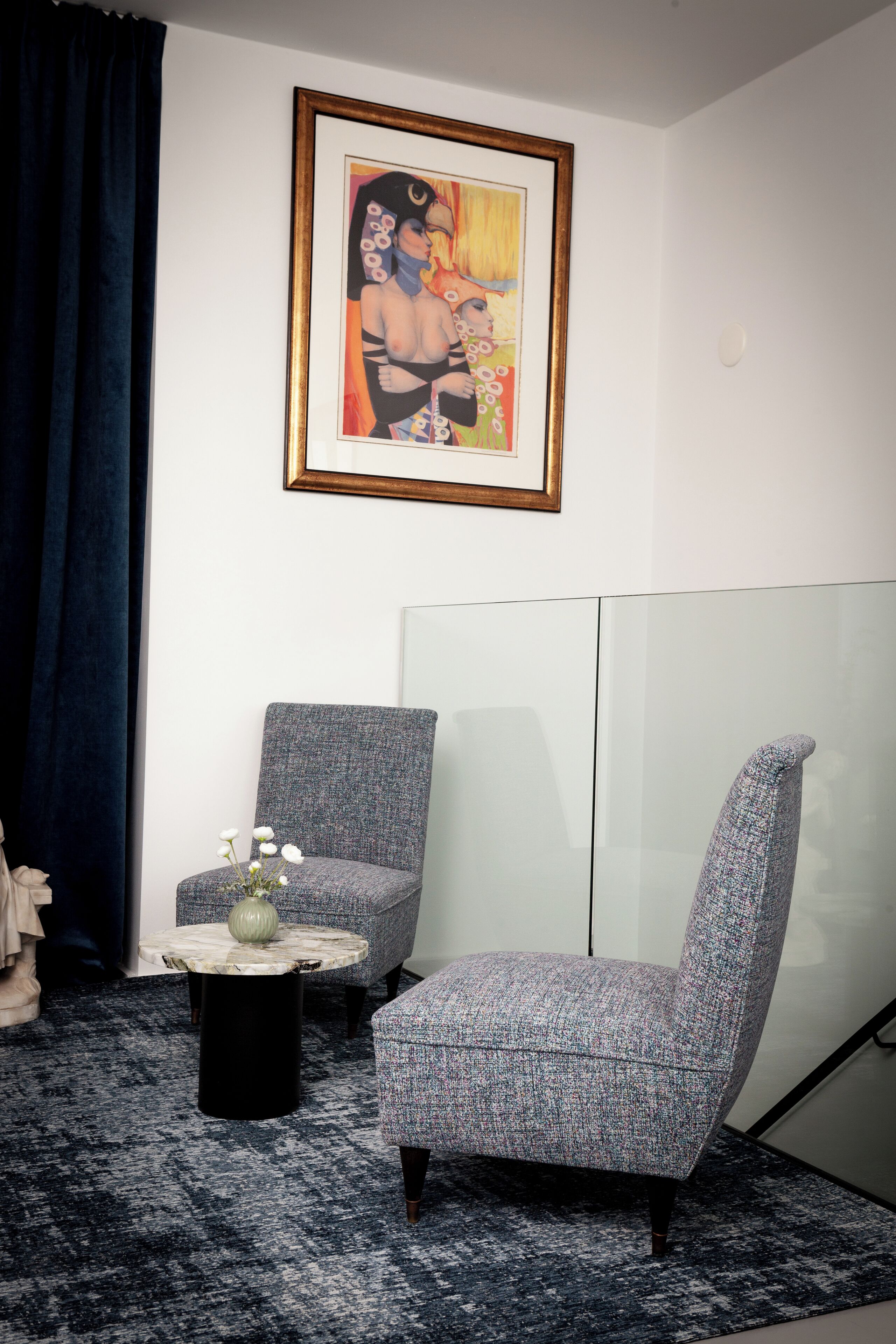 Quarto com poltrona cinzenta, mesa redonda pequena com um vaso, e uma obra de arte emoldurada na parede.