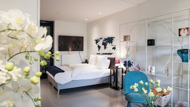 A modern bedroom with a bed, a blue chair, a world map on the wall, and a glass table with a vase of flowers. - Suite Aux Anges (Antwerp)