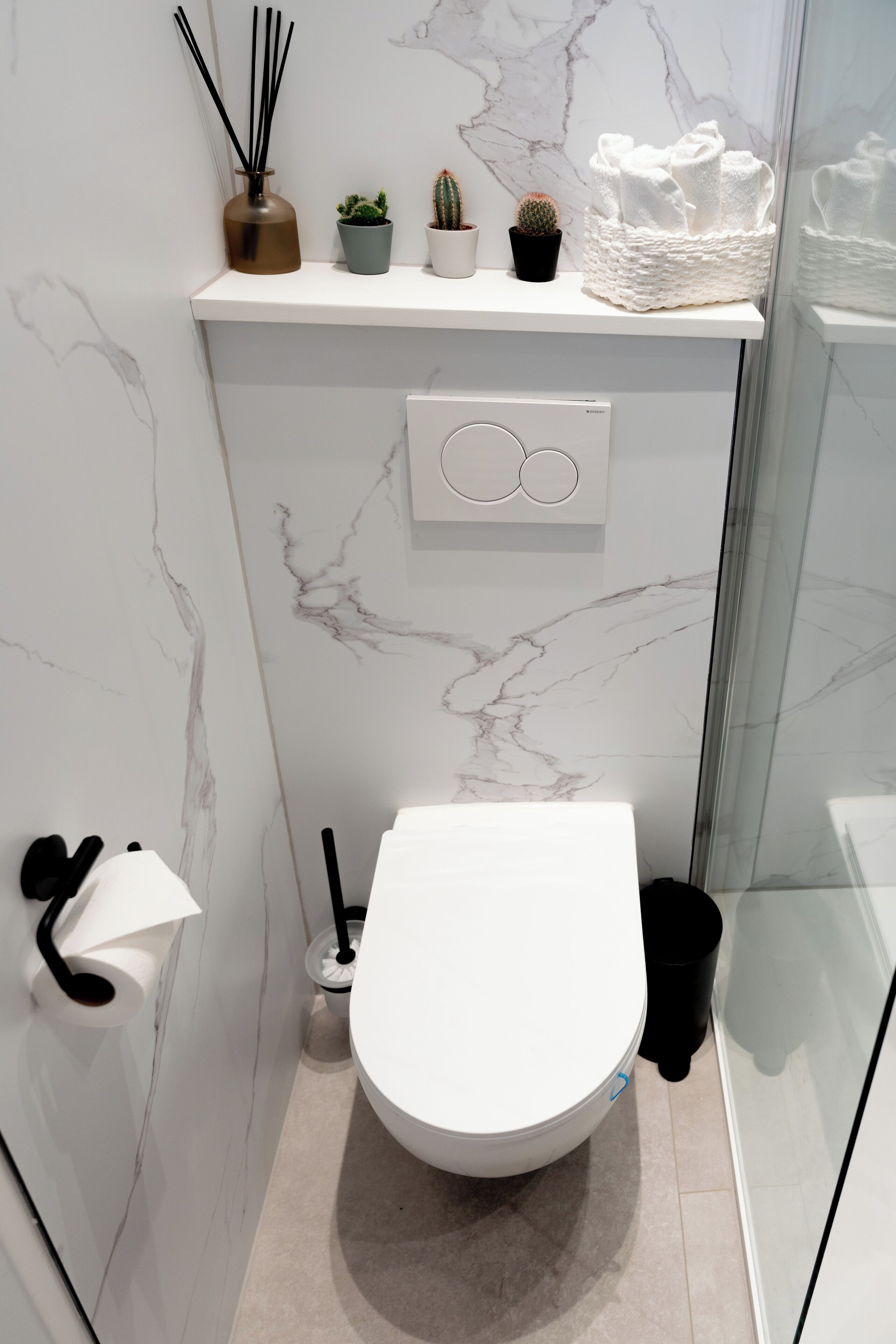 A bathroom with a white toilet, a marble wall, a small shelf with plants, and a white towel basket.