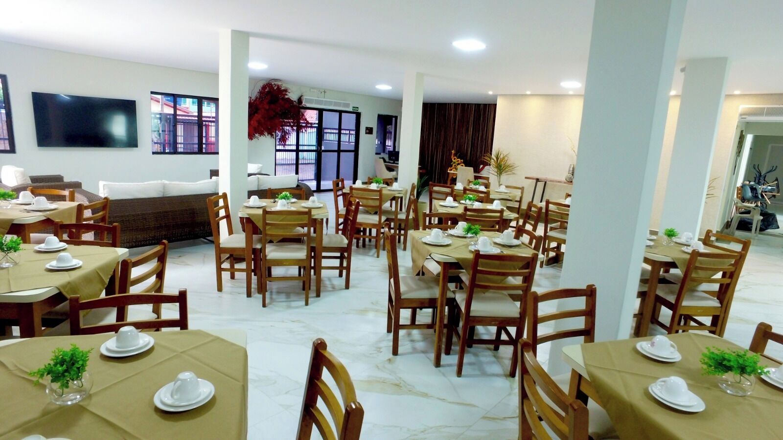 A spacious dining area with multiple tables and chairs, a large window with a view, and a flat-screen TV mounted on the wall.