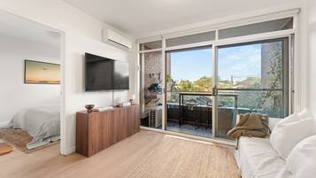 A modern living room with a flat-screen TV, a sofa, a dining area with a table and chairs, and a large sliding glass door leading to an outdoor balcony.