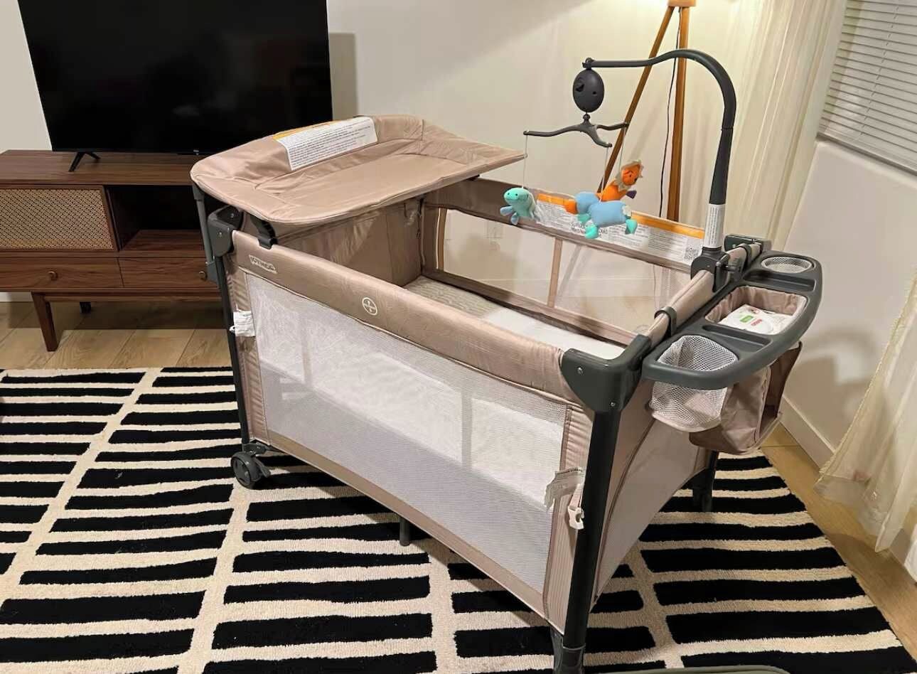 A baby crib with a mesh side and a canopy, positioned on a black and white striped rug in a room with a wooden cabinet and a television in the background.