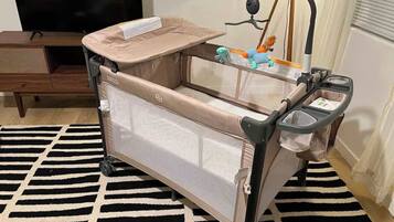 A baby crib with a mesh side and a canopy, positioned on a black and white striped rug in a room with a wooden cabinet and a television in the background.