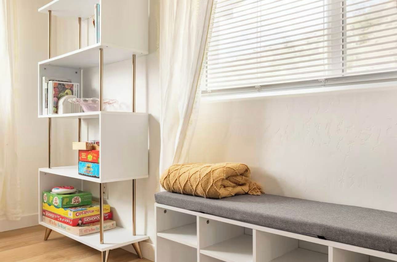 Um quarto com uma estante branca, um banco cinza e uma janela com persianas.