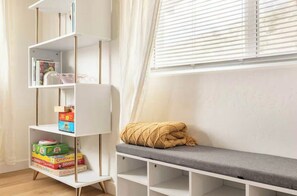 A room with a white bookshelf, a grey bench, and a window with blinds.