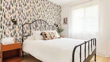 Una cama bien hecha con edredón blanco y almohadas estampadas, una mesita de noche con lámpara y un papel tapiz decorado.