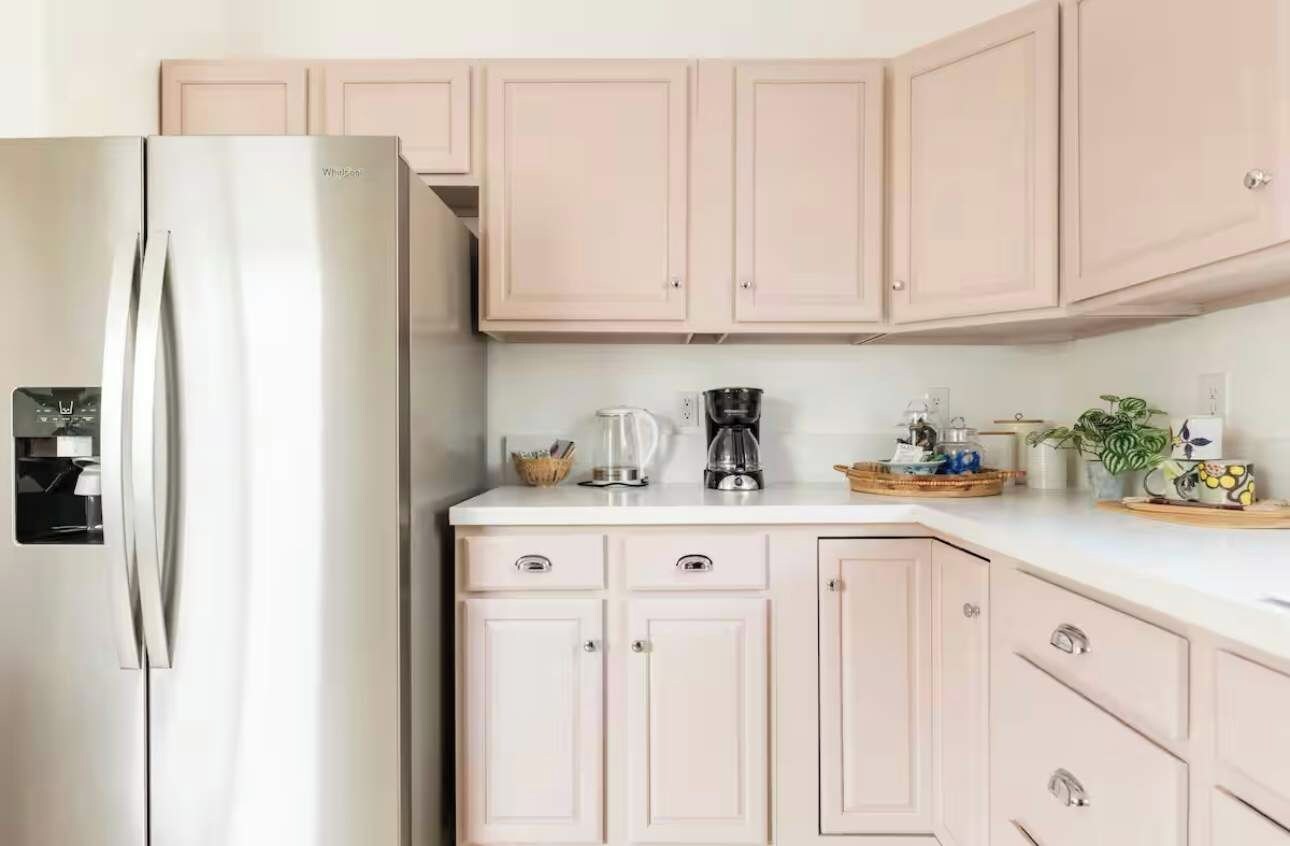 A kitchen with light pink cabinets, a stainless steel refrigerator, a coffee maker, and a kettle on the counter.