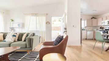 A modern living room with a sofa, a coffee table, and a chair. There is a kitchen area with cabinets and a dining table.