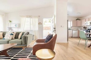 A modern living room with a sofa, a coffee table, and a chair. There is a kitchen area with cabinets and a dining table.