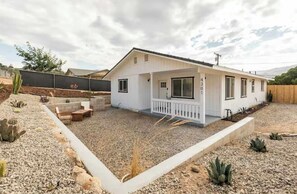 Une maison blanche d’un seul étage, avec un porche à l’avant, une petite terrasse et une allée gravillonnée.