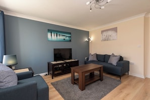 A modern living room with a dark blue sofa, a wooden coffee table, a TV on a stand, and artwork on the walls. - Toast Stays - Hillbrook (Thornaby)