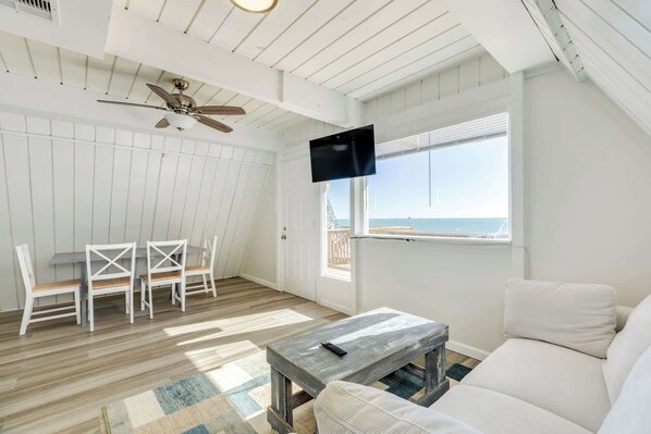 A room with a ceiling fan, a TV mounted on the wall, a sofa, a table, and a view of the ocean through a window. - Unique Beachfront Escape Directly Over The Ocean (Surfside Beach)