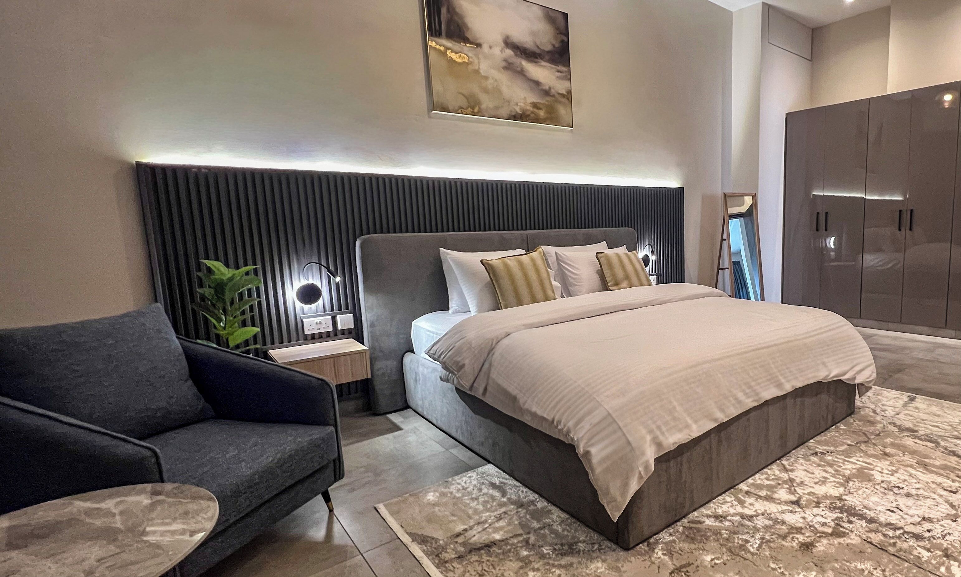 A modern bedroom with a large bed, a dark armchair, a bedside table with a lamp, and a wardrobe.