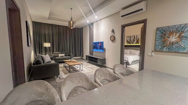 A modern living room with a sofa, coffee table, and TV. There is a dining area with chairs and a table. The room has a large window with curtains, an air conditioning unit, and a painting on the wall. - Anthonian Suites (Accra)